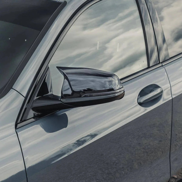 The side of a sleek car features a shiny side mirror reflecting clouds. The car's metallic surface also shows a mirror-like reflection, set outdoors under a bright sky.