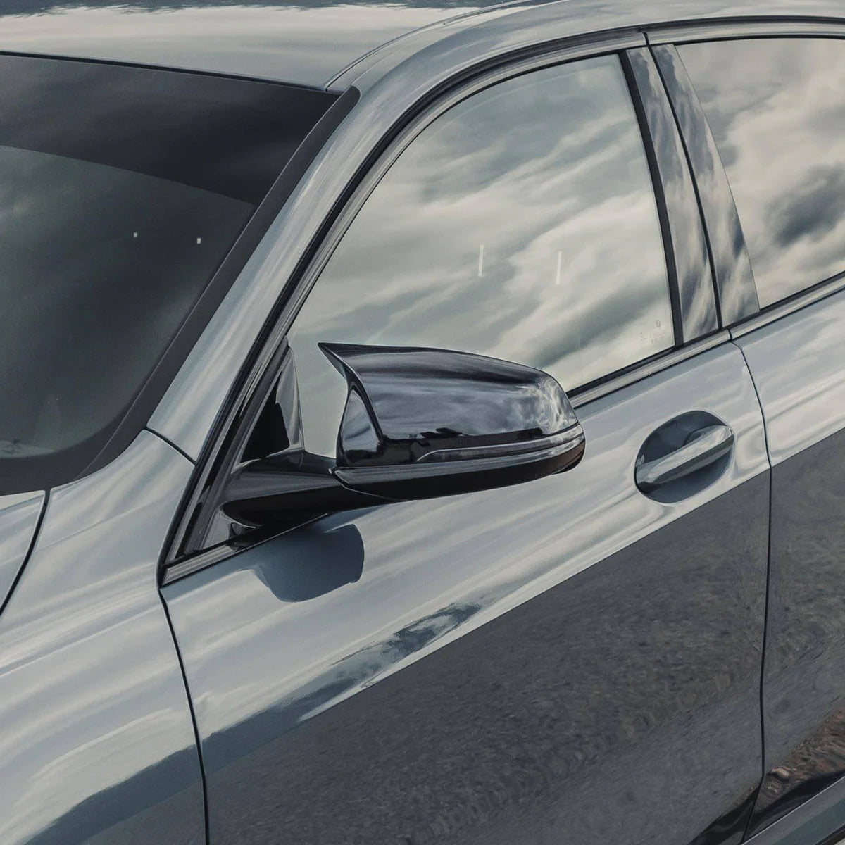 Car side mirror reflects sky and clouds, mounted on a sleek blue vehicle in an outdoor setting. The car door and handle are visible, highlighting modern automotive design.