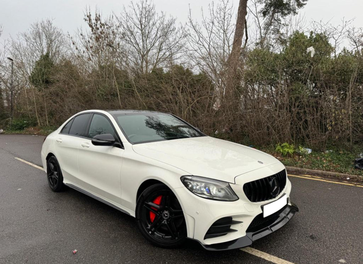 A white car with black rims is parked on a wet road beside leafless trees and bushes. The vehicle features a sleek design with red brake calipers.