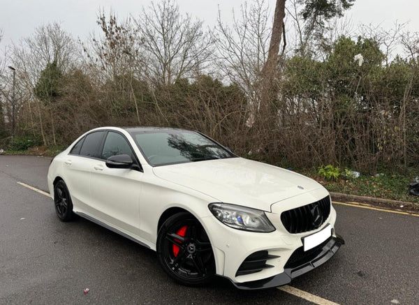 A white car with black rims is parked on a wet road beside leafless trees and bushes. The vehicle features a sleek design with red brake calipers.