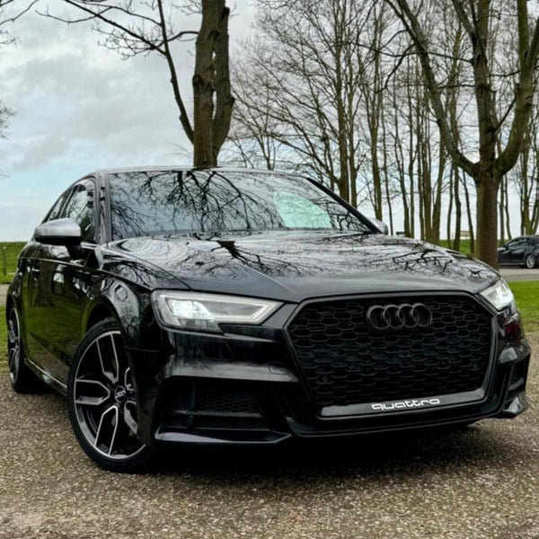 A black Audi car is parked on a concrete surface, surrounded by bare trees. The front grille features the Audi logo and the word "quattro."