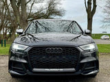 A black Audi car stands parked on a textured surface with trees and grass surrounding it. The front grille prominently displays the Audi logo and 