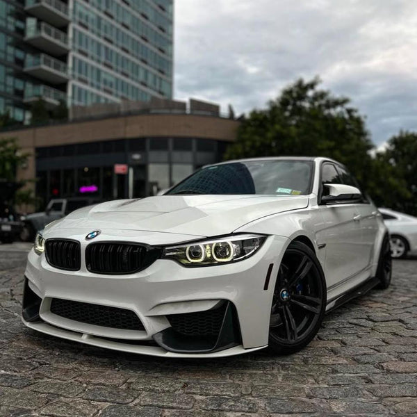 A white BMW car is parked on a cobblestone street, with distinct LED headlights on and a modern building in the background. Trees and another vehicle complete the urban setting.