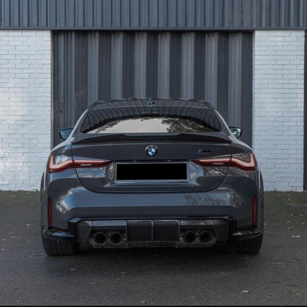 A gray BMW car parked in front of a white brick wall with a dark metal door. The car features visible dual exhausts and taillights.