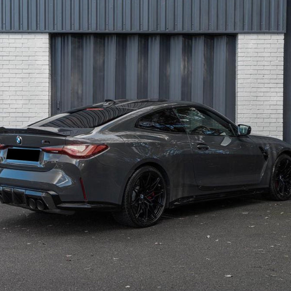 A gray sports car is parked on an asphalt surface in front of a corrugated metal building with a white brick section.