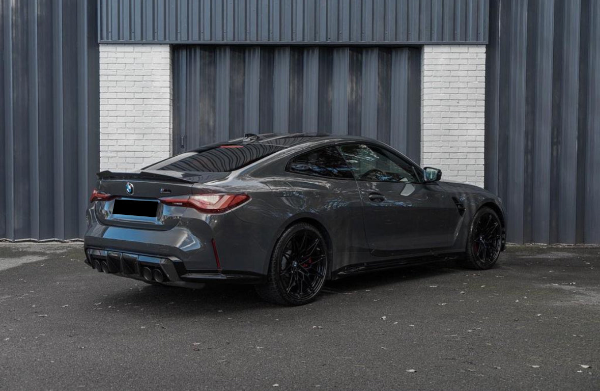 A gray sports car is parked on an asphalt surface in front of a corrugated metal building with a white brick section.