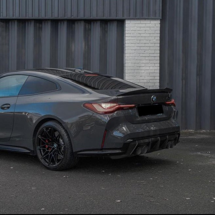 A sleek dark gray sports car is parked on a paved surface, facing away. It is positioned against a backdrop of dark corrugated metal and a section of white brick wall.