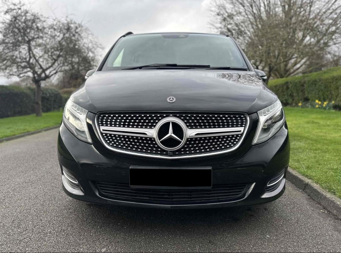 A black Mercedes-Benz van is parked on a paved road, surrounded by green grass and trees, under an overcast sky. The prominent logo and grille are visible.