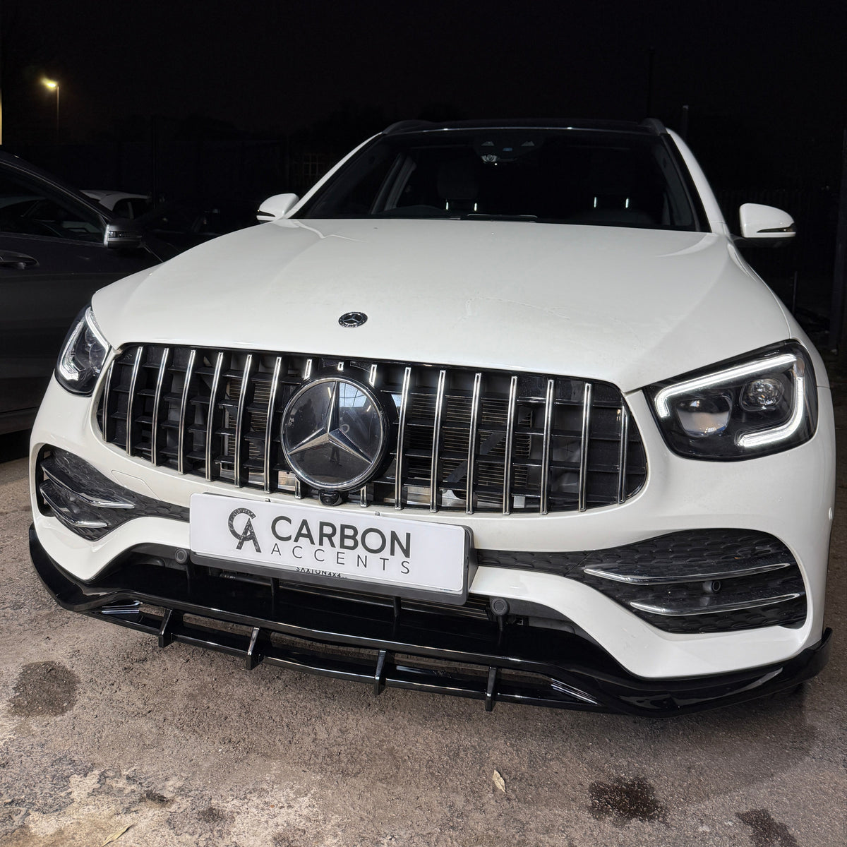 A white luxury car with a prominent emblem and grille, parked in a dimly lit area. The front license plate reads "CARBON ACCENTS."