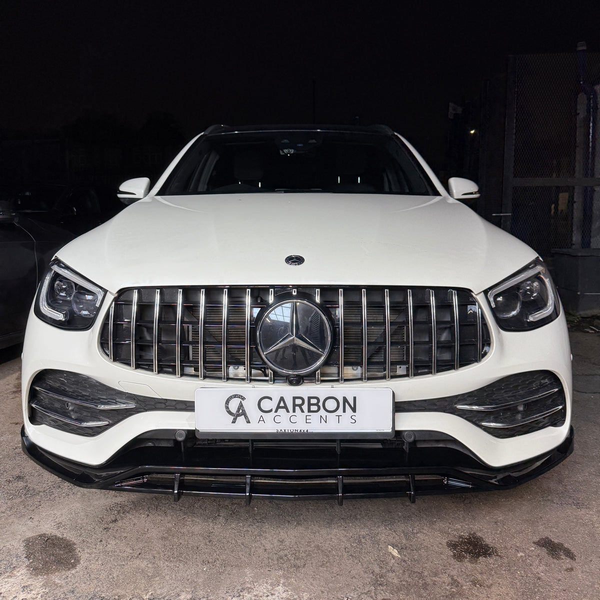 A white SUV is parked on a concrete surface in front of a dark backdrop. The front grille features a prominent Mercedes emblem. The license plate reads "CARBON ACCENTS".