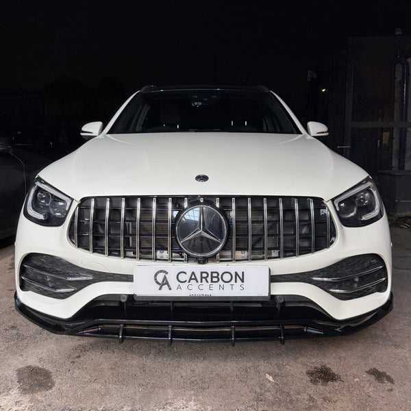 A white SUV is parked on a concrete surface in front of a dark backdrop. The front grille features a prominent Mercedes emblem. The license plate reads "CARBON ACCENTS".