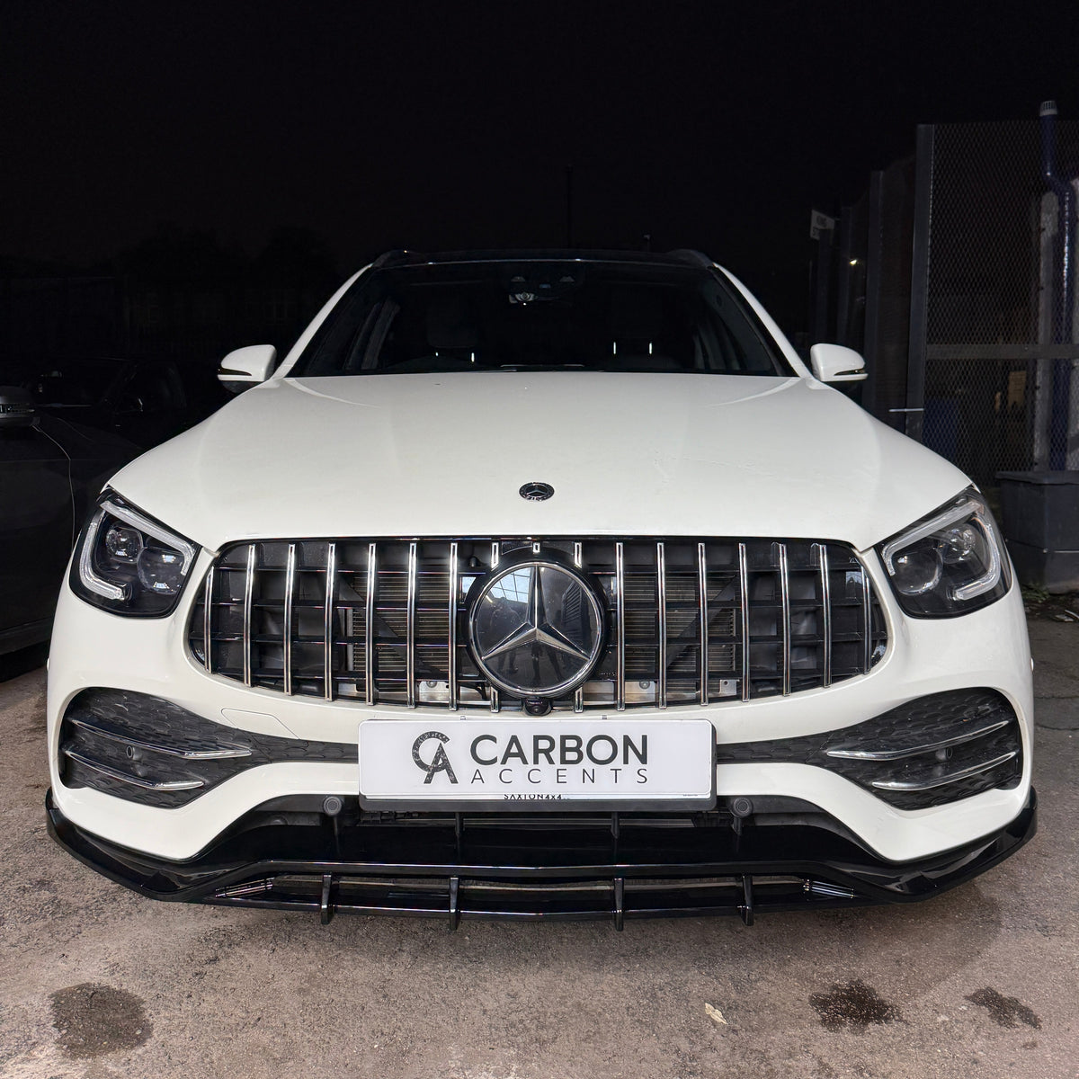 A white SUV with a prominent front grille bears a badge and is parked in a dimly lit area. The license plate reads "CARBON ACCENTS AUTOMOTIVE."