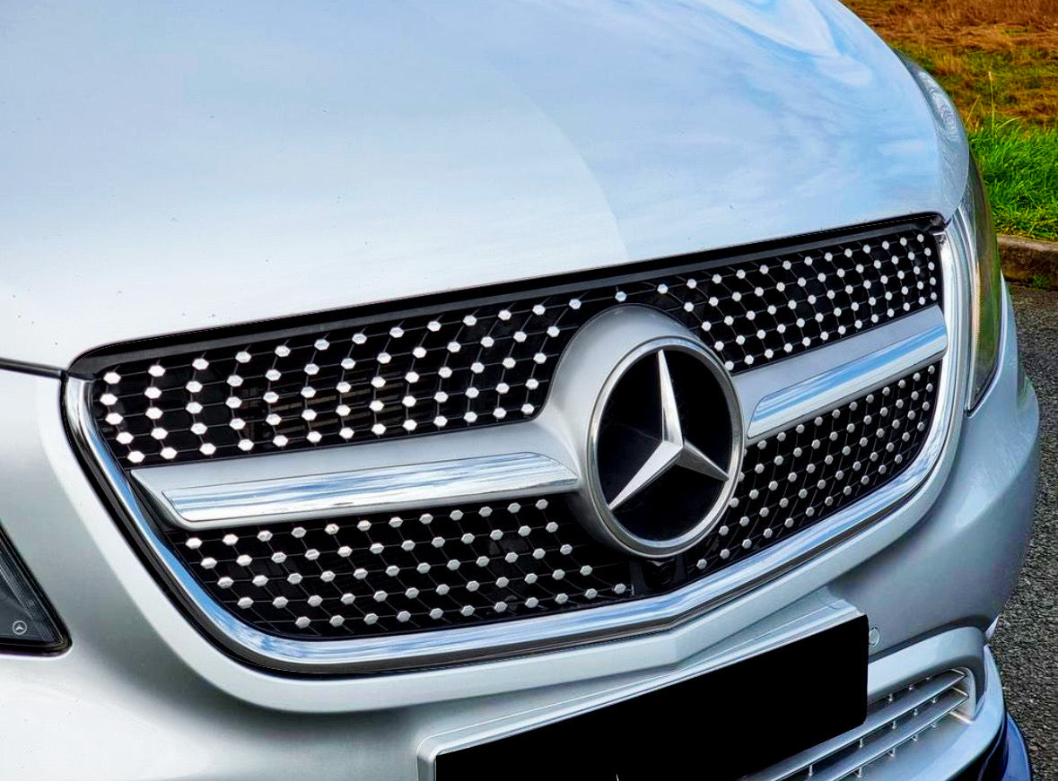 A silver car grille displays a circular emblem with a three-point star, surrounded by a pattern of dots. It's parked on an asphalt road near green grass.
