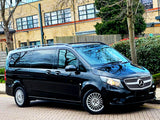 A black Mercedes-Benz van is parked on a paved street. The setting includes a background with a brick building, trees, and a hedge.