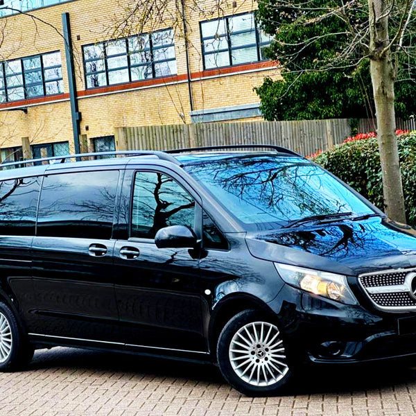 A black Mercedes-Benz van is parked on a paved street. The setting includes a background with a brick building, trees, and a hedge.