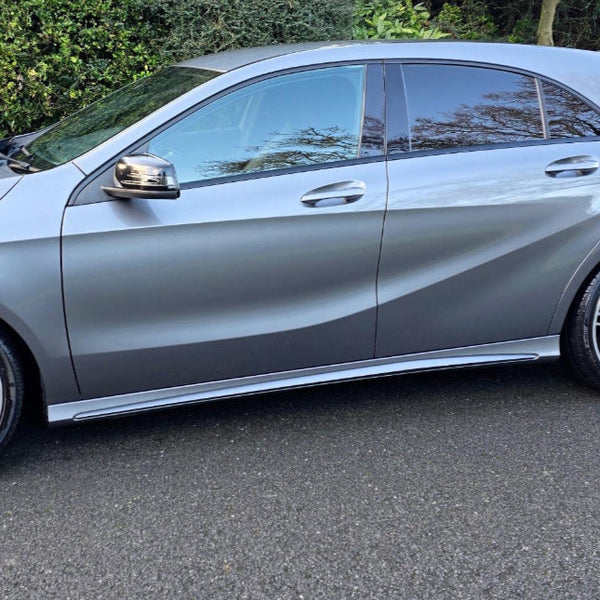 A silver car is parked on a road, surrounded by greenery. The vehicle's side profile and closed windows are visible.