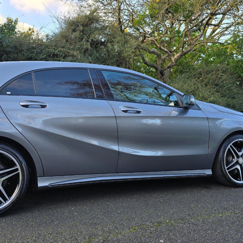 A silver car parked on a road; surrounded by lush, green trees and bushes. The car features tinted windows and black alloy wheels, under a partly cloudy sky.
