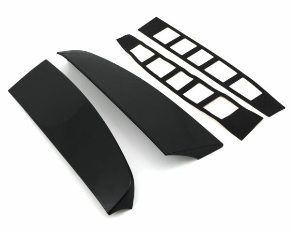 Two glossy black car spoiler pieces lay parallel, accompanied by two sets of adhesive strips with rectangular cutouts, all set against a plain white background.