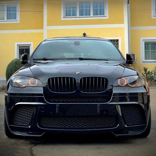 A black BMW with illuminated circular headlights is parked in front of a yellow building with white trim and windows, creating a striking contrast between the car and its surroundings.