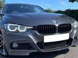 A silver BMW car is parked on a paved surface, with its headlights on. The background features greenery and a clear blue sky.