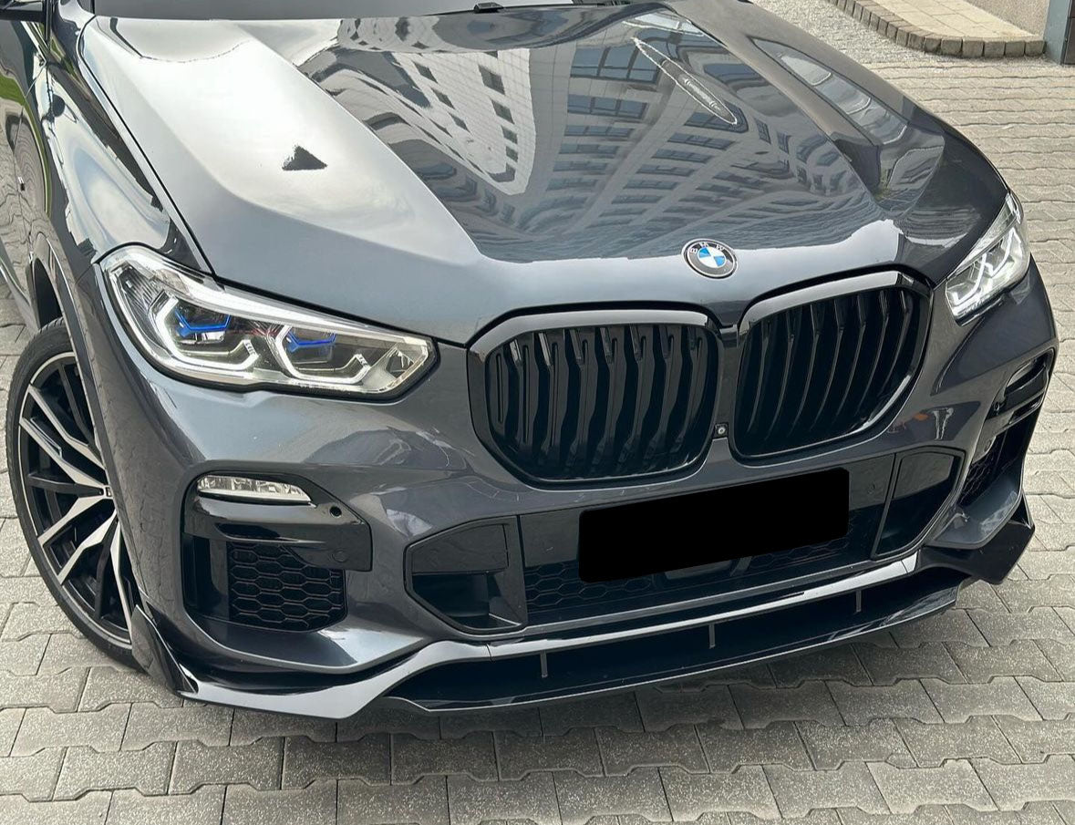 A sleek, gray car with a prominent grille and shiny, intricate headlights is parked on a patterned concrete surface, reflecting nearby buildings on its glossy hood.