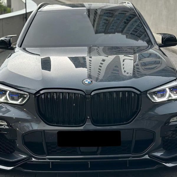 Car parked on a paved area. The sleek, black vehicle has prominent grille and headlights, with reflections of tall buildings on its glossy surface. The environment appears urban and modern.