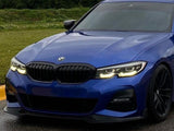 A blue BMW with its headlights on is parked on a wet road, surrounded by grass and trees in the background.
