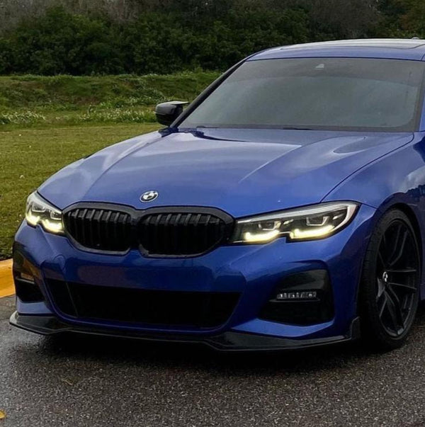 A blue BMW with its headlights on is parked on a wet road, surrounded by grass and trees in the background.