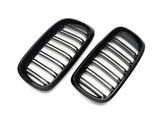 Two black, rectangular car grilles with vertical slats rest diagonally against a plain white background, emphasizing their sleek, glossy design.