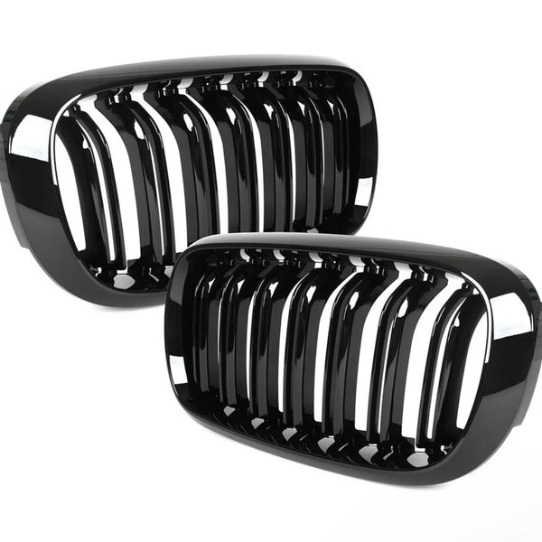 Two black, glossy car grilles with vertical slats lean against each other, reflecting light. They are set against a plain white background.