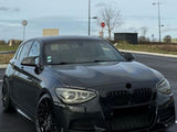 A dark, sleek car is parked on an asphalt road, surrounded by sparse trees and traffic signs under a cloudy sky.
