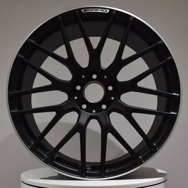 A black alloy wheel with multiple spokes, labeled "AMG" at the top, is resting upright against a neutral background atop a white circular platform.