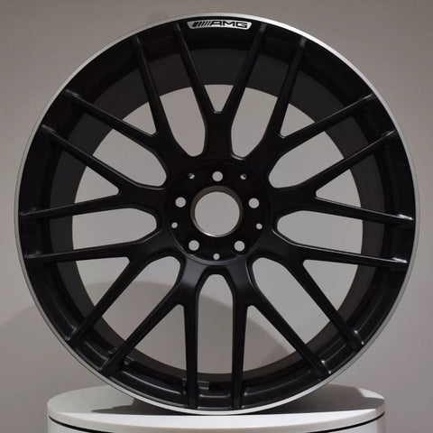 A black alloy wheel with multiple spokes, labeled "AMG" at the top, is resting upright against a neutral background atop a white circular platform.