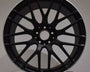A black, multi-spoke AMG car rim is displayed upright against a neutral background, resting on a white surface.