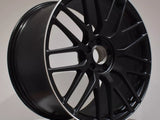 A black alloy wheel with intricate spoke design is displayed on a white platform. The rim features a silver edge, and 