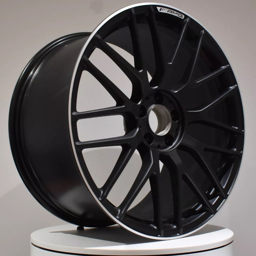 A black alloy wheel displayed upright. Its multi-spoke design features "AMG" text on one spoke. It is positioned on a white circular platform against a plain gray background.