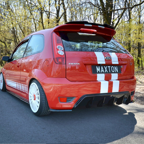 A red Ford Fiesta with white stripes is stationary on a tree-lined road. The license plate reads "MAXTON."