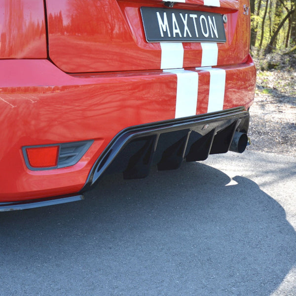 A red car's rear displaying a black diffuser and dual exhaust pipes, parked on a paved road with trees. The license plate reads "MAXTON," and white stripes adorn the vehicle.