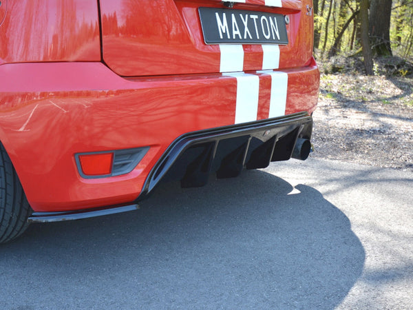 A red car's rear displaying a black diffuser and dual exhaust pipes, parked on a paved road with trees. The license plate reads "MAXTON," and white stripes adorn the vehicle.