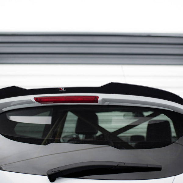 A white car's rear with a black spoiler and rear window wiper is pictured against a garage door. The spoiler has a small logo near the middle top.
