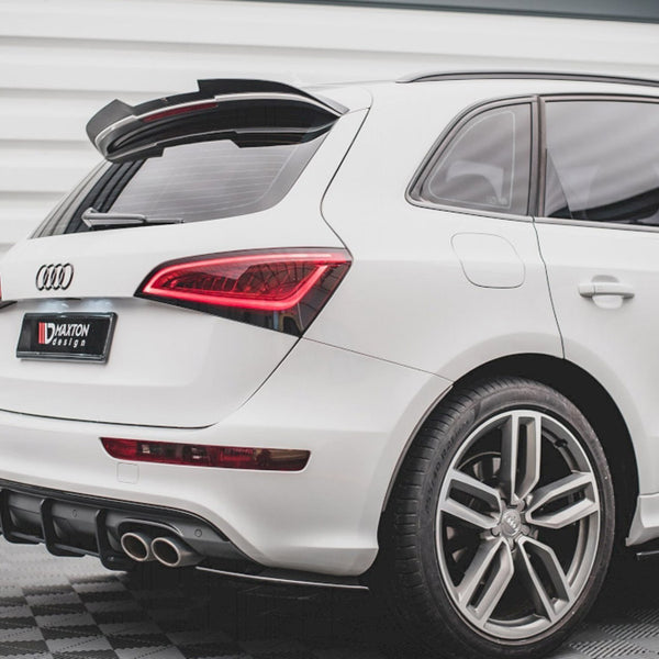 A white Audi SUV is parked on a checkered floor, featuring a prominent rear spoiler and dual exhaust pipes. Text on a license plate reads "Maxton Design."