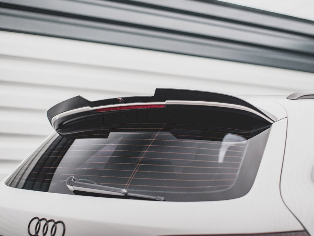 A white car's rear, featuring a roof spoiler and rear windshield wiper, is parked outside with a garage door in the background. The Audi logo is visible at the bottom.