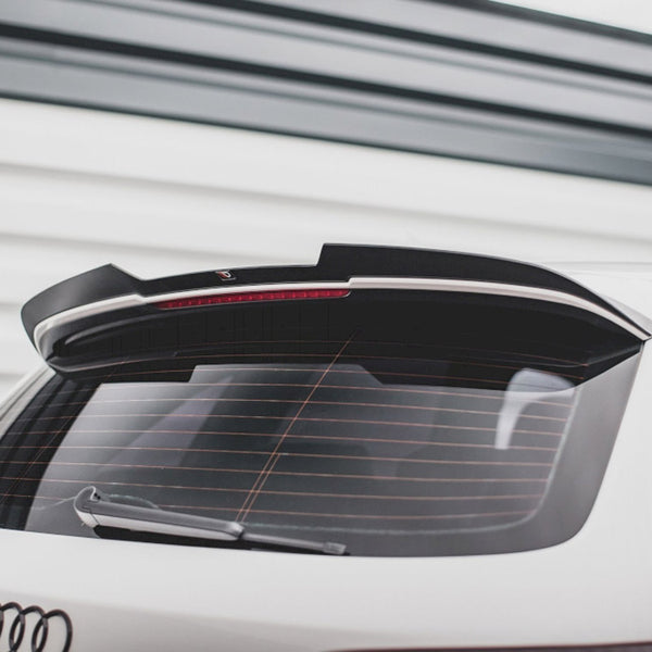 A white car's rear, featuring a roof spoiler and rear windshield wiper, is parked outside with a garage door in the background. The Audi logo is visible at the bottom.