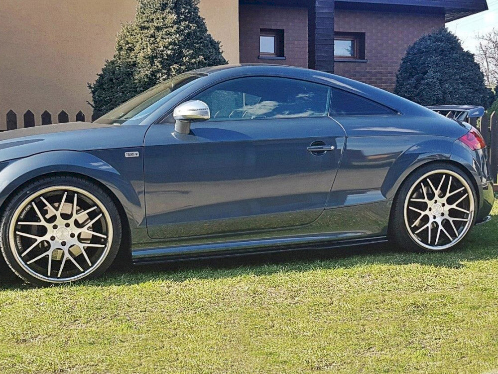 A sleek, dark gray sports car with large alloy wheels is parked on a grassy lawn in front of a house, bordered by evergreen shrubs and a wooden fence.