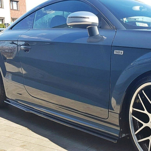A dark blue sports car parked on a sunlit driveway, featuring large black alloy wheels with silver spokes and a "Traffic W" badge on its side.
