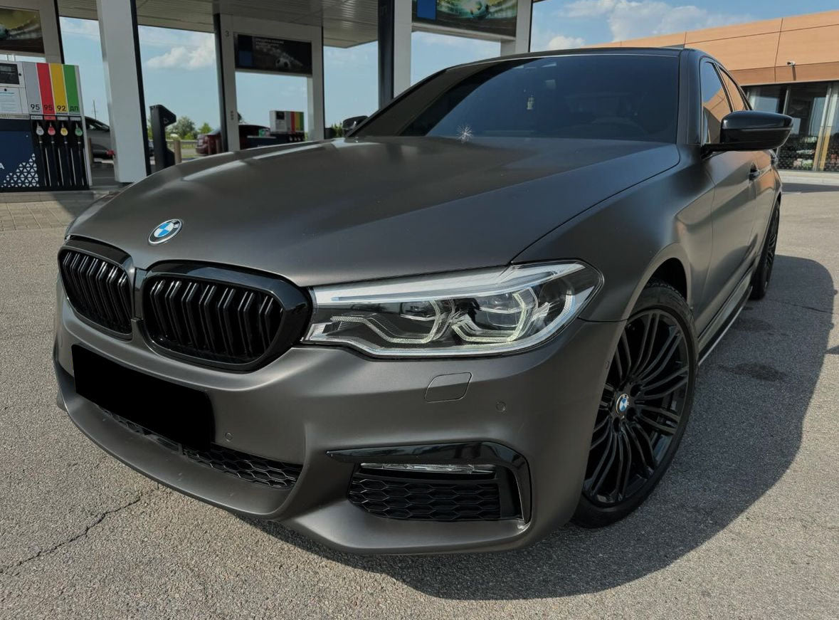 A sleek, matte gray BMW sedan is parked at a gas station with fuel pumps visible in the background under a clear sky.
