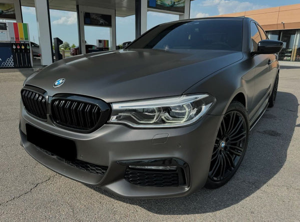 A sleek, matte gray BMW sedan is parked at a gas station with fuel pumps visible in the background under a clear sky.