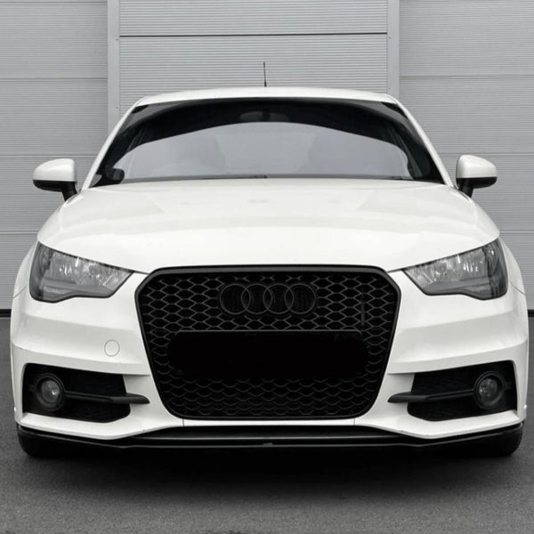 A white car with a black honeycomb grille and Audi emblem is stationary, displayed front-facing against a light gray, vertically paneled wall in a parking area.