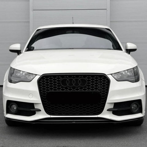 A white car with a black honeycomb grille and Audi emblem is stationary, displayed front-facing against a light gray, vertically paneled wall in a parking area.