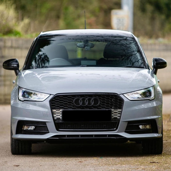 A silver car with a black grille and Audi logo is parked on a road, surrounded by blurred greenery and a brick wall.